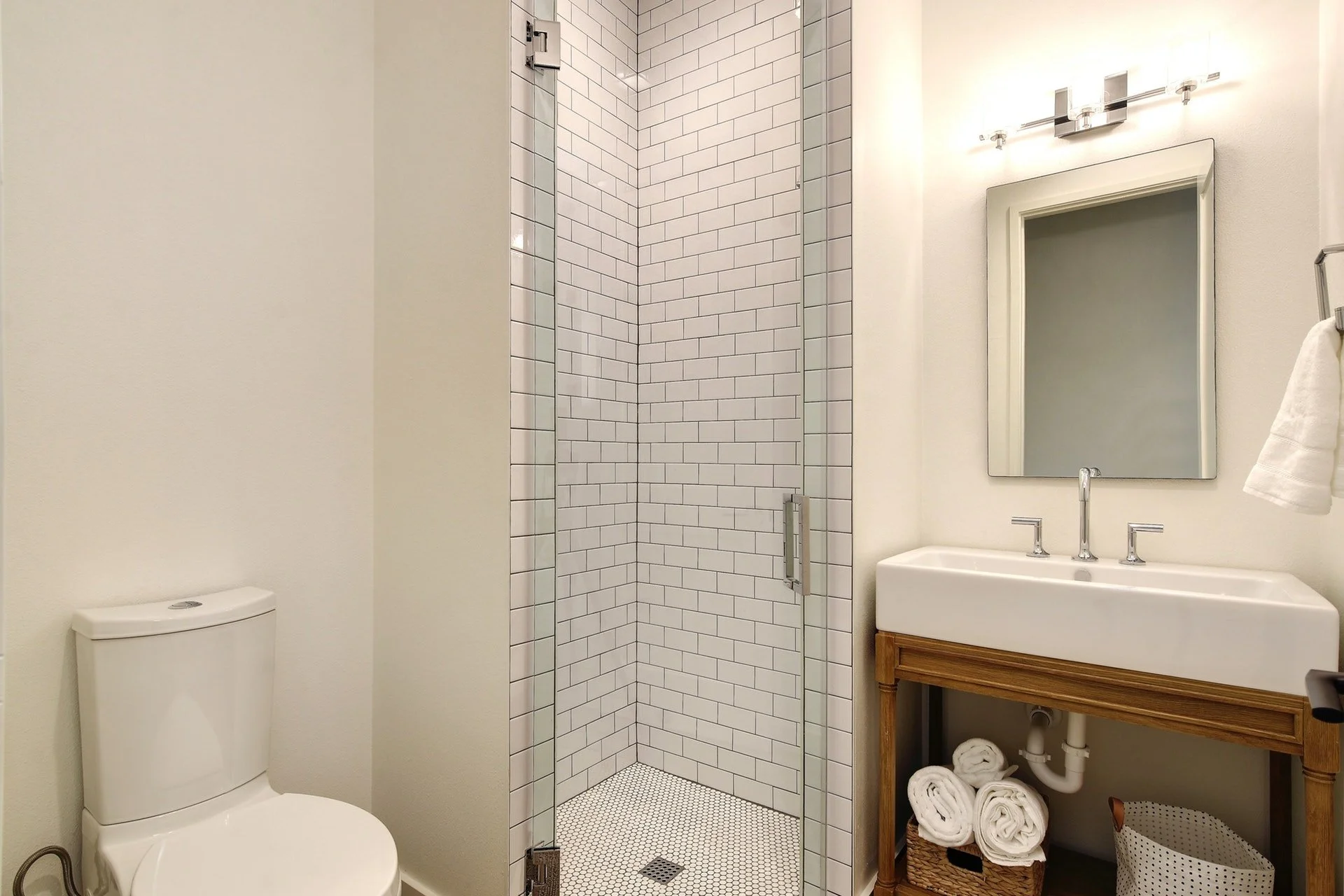 Modern bathroom with a white toilet, a shower with white subway tiles, a vanity with a mirror and a towel rack, and rolled towels in a basket underneath.