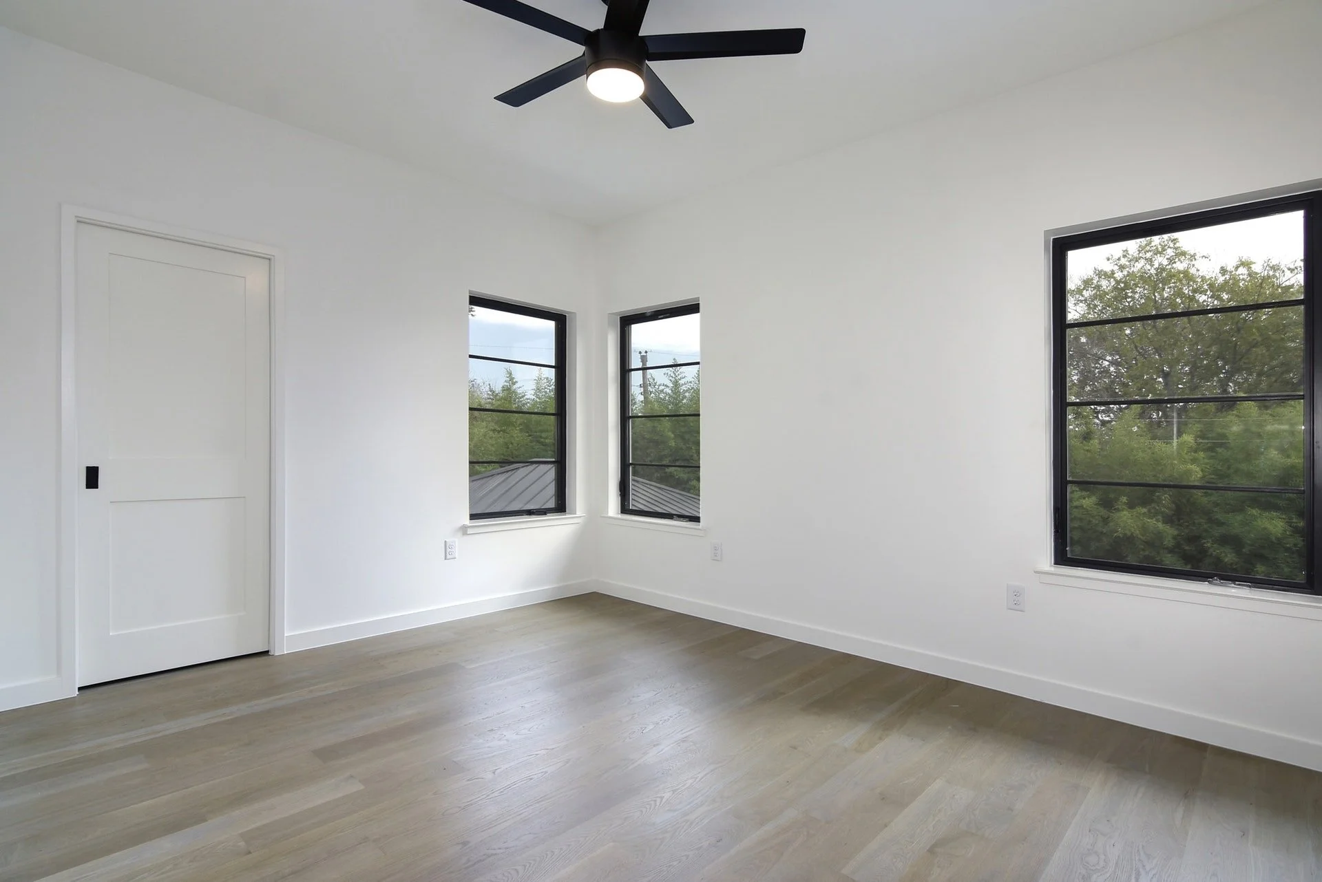 Empty room with white walls, light wooden floor, black ceiling fan, and three large windows showing trees and cloudy sky outside.