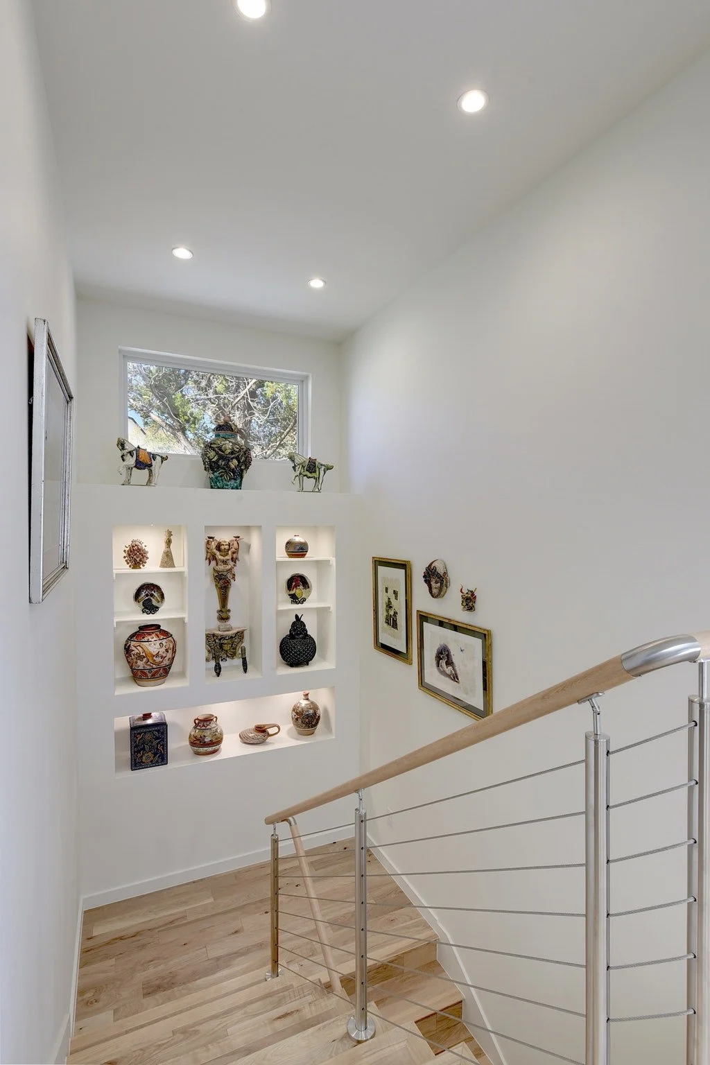 Interior view of a staircase landing with a white wall display shelf filled with decorative pottery and sculptures, framed artwork on the wall, a large window at the top, and a metal handrail on the staircase.