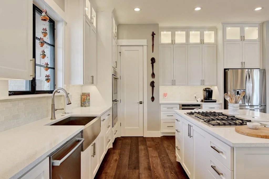 Modern white kitchen with stainless steel appliances, wooden floor, and decorative wall hanging.