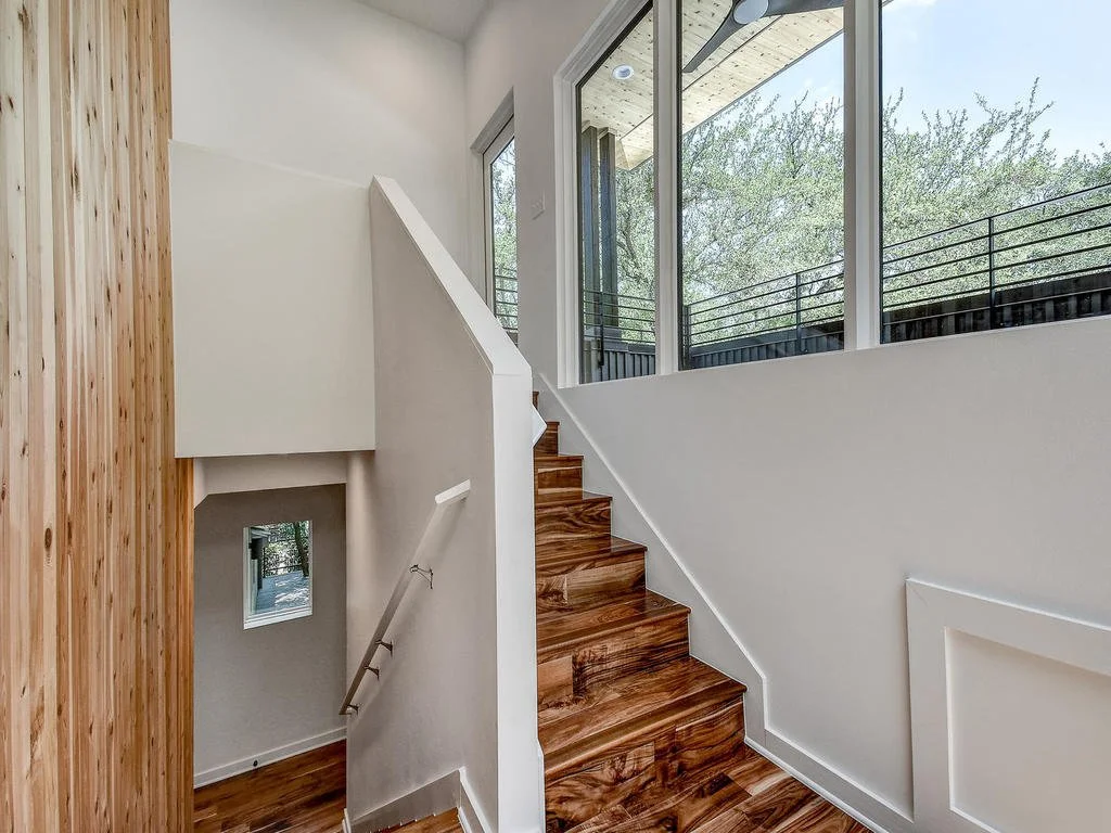 Interior view of a staircase with wooden steps, white walls, large windows, and a balcony railing outside.