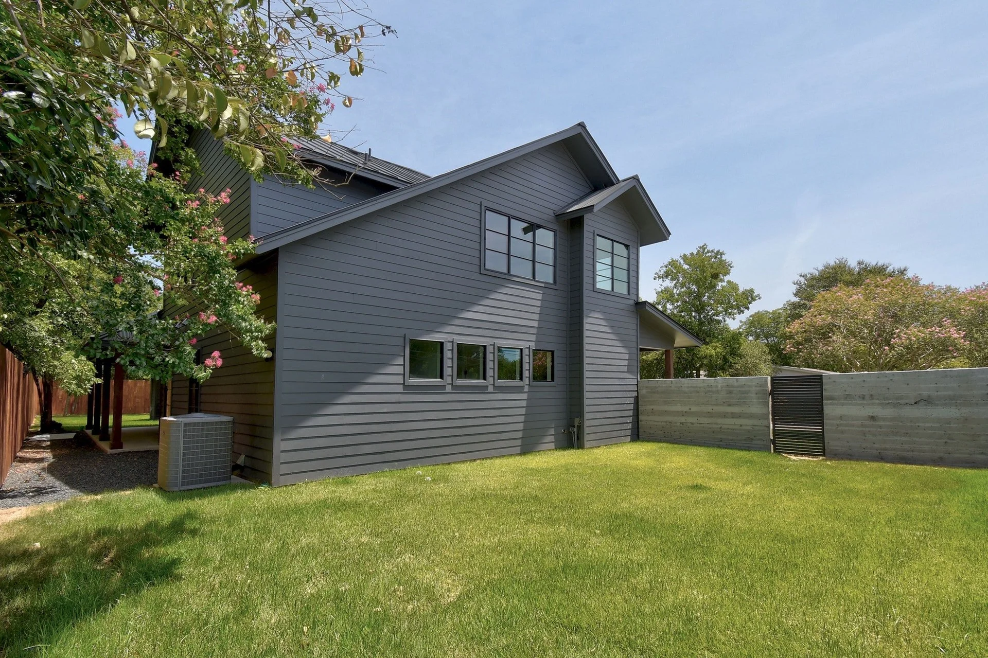 Modern gray house with multiple windows, surrounded by green lawn and trees with pink and white blossoms, under a blue sky.