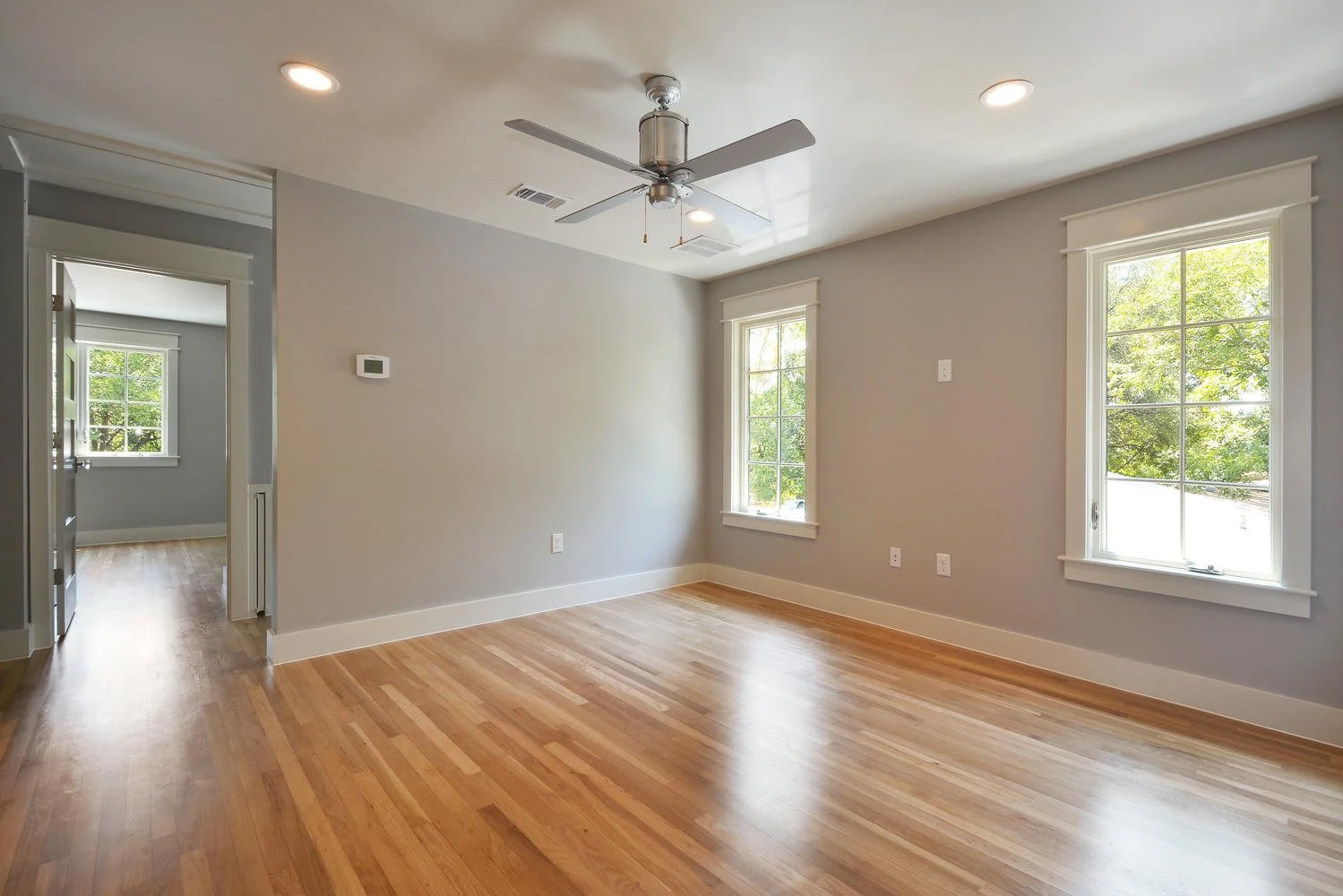 Empty room with hardwood floors, two windows, a ceiling fan, and neutral-colored walls.
