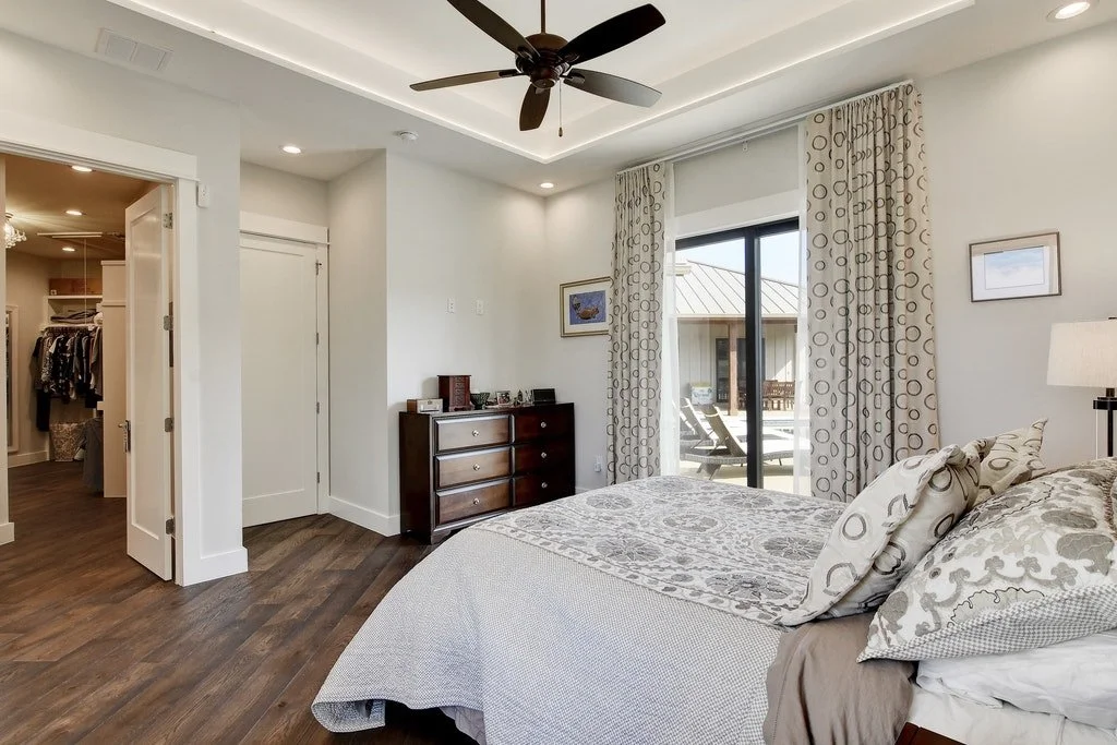 A bedroom featuring a bed with patterned pillows and bedding, a sliding glass door leading to an outdoor patio with chairs, a dresser with decorative items, a ceiling fan, and a walk-in closet with clothes. The room has white walls, wooden flooring, 