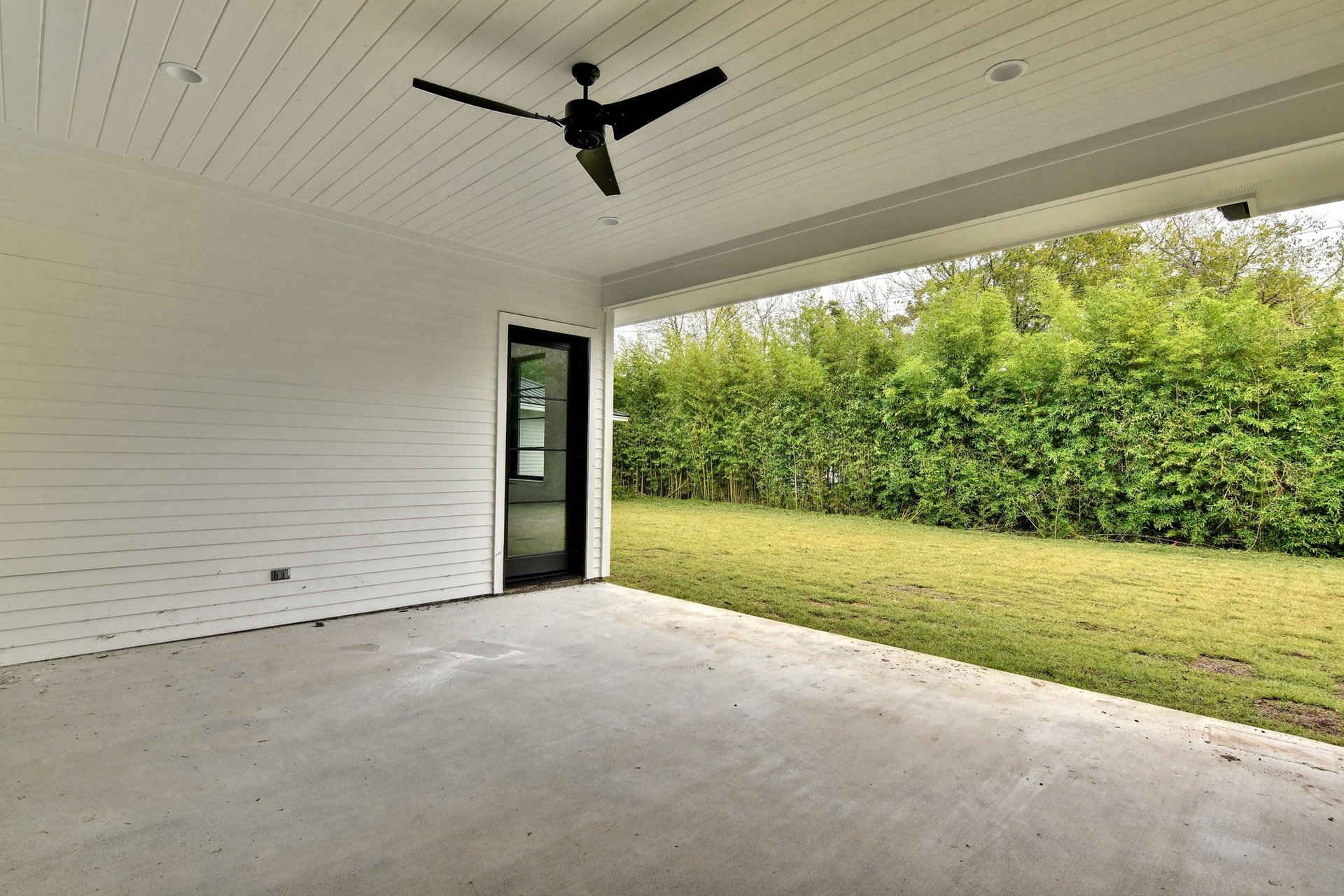 View of a covered patio with a white ceiling, black ceiling fan, and sliding glass door leading into a house. The patio has a concrete floor and overlooks a grassy yard and green trees in the background.