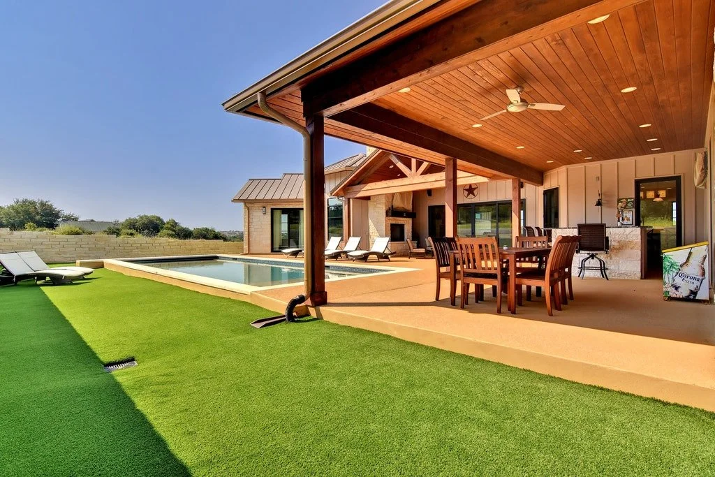 A backyard patio with a swimming pool, outdoor dining table with chairs, lounge chairs, and a covered seating area with a fireplace, ceiling fan, and bar area, lush green lawn, and clear blue sky.