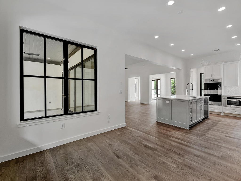 Empty, modern open-concept kitchen and living area with white cabinets, a large kitchen island, hardwood floors, and black-framed windows.
