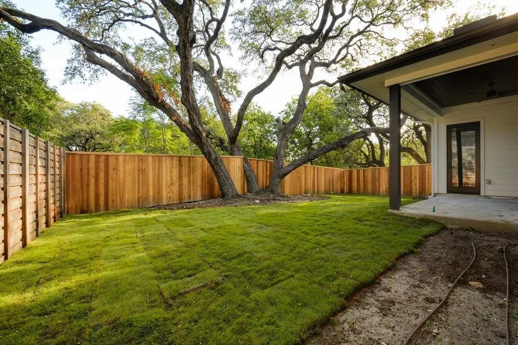 A backyard with a lush green lawn, a large tree with sprawling branches, a wooden privacy fence, and part of a house with a covered patio area.