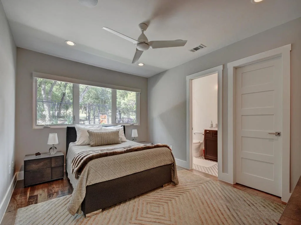 Bedroom with large window, bed with pillows, nightstands with lamps, ceiling fan, and an en-suite bathroom.