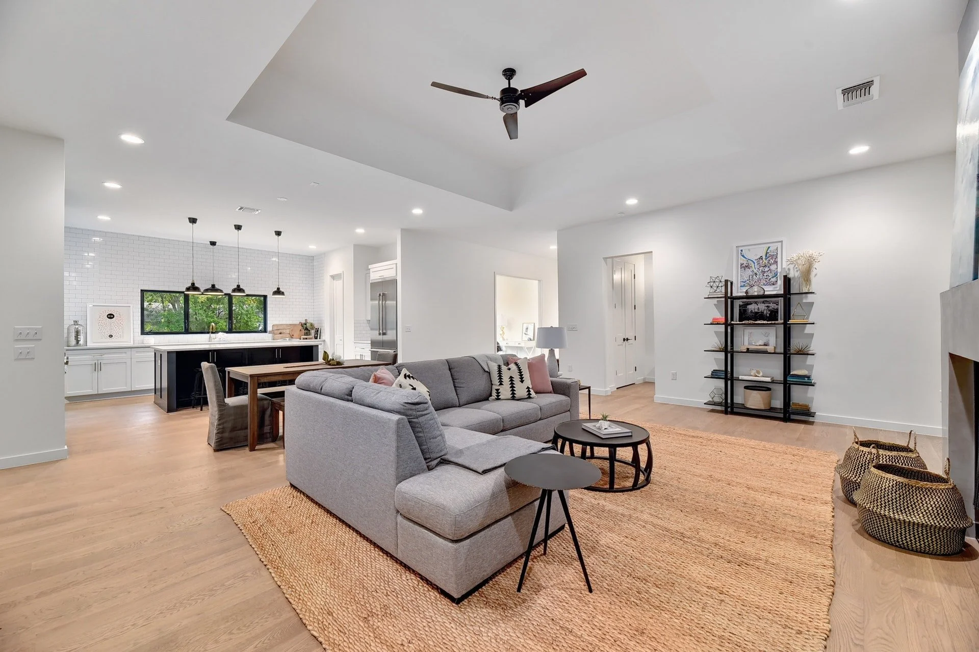 Open-concept living room with gray sectional sofa, black round coffee tables, beige rug, and a black metal bookshelf. The space features a white ceiling with recessed lighting, a ceiling fan, and a view into the kitchen with white cabinetry, black is