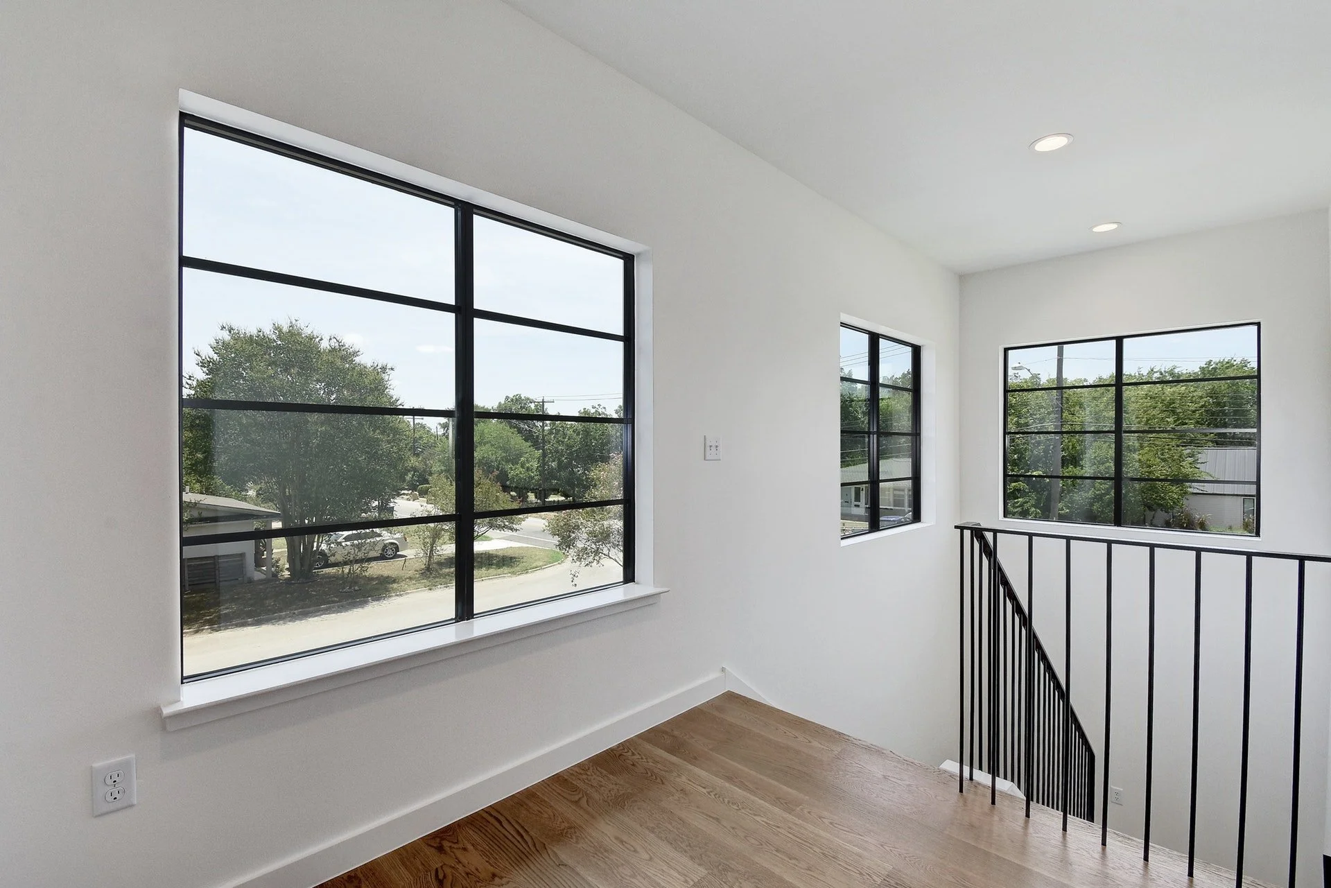 Interior view of a modern stairway landing with large windows showing trees and a street outside, white walls, hardwood flooring, and a black metal railing.