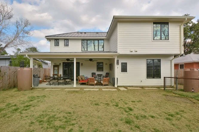 A two-story house with a spacious backyard, covered patio with outdoor furniture, and a lawn with patches of dirt, enclosed by a wooden fence.