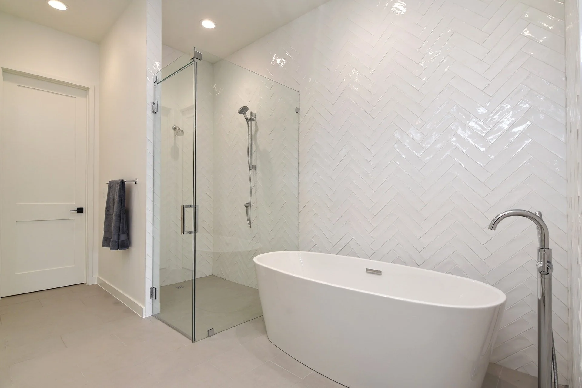 Modern bathroom with white herringbone tile wall, bathtub, glass-enclosed shower with handheld shower on wall, white door, gray towel on wall, and brushed nickel faucet.