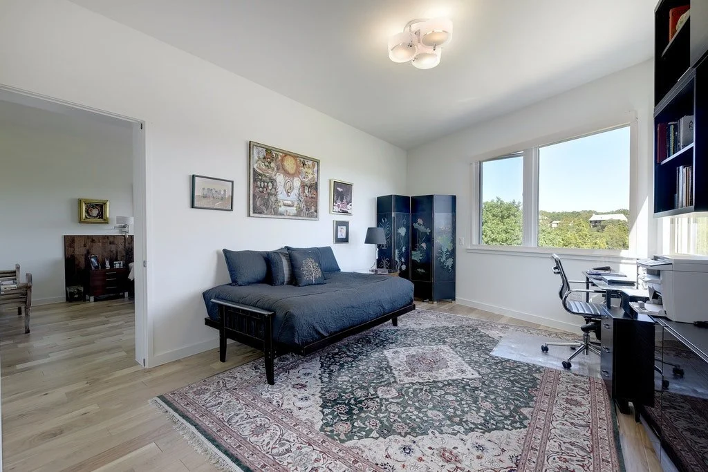 A well-lit living room with a large window, a black daybed with pillows, a patterned rug, a black folding screen, a desk with a printer and chair, and a bookshelf. A doorway shows a bedroom with a bed and nightstand.