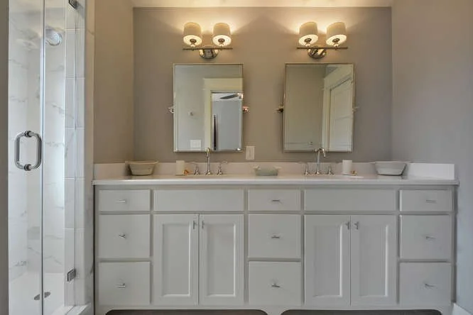 A bathroom double vanity with two mirrors, four light fixtures, and a white countertop with cabinets below. There are soap dispensers and bowls on the counter, and a glass shower door is visible on the left.