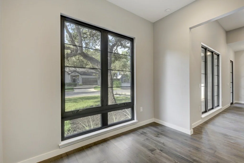 Empty room with large windows overlooking a tree and street, hardwood floors, and white walls.