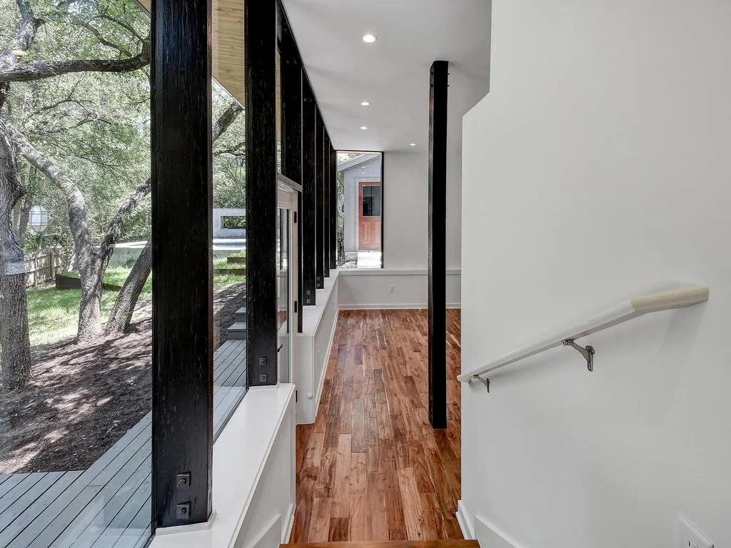 Interior view of a modern hallway with wooden floors, large windows showing trees outside, a white wall, and a staircase handrail.