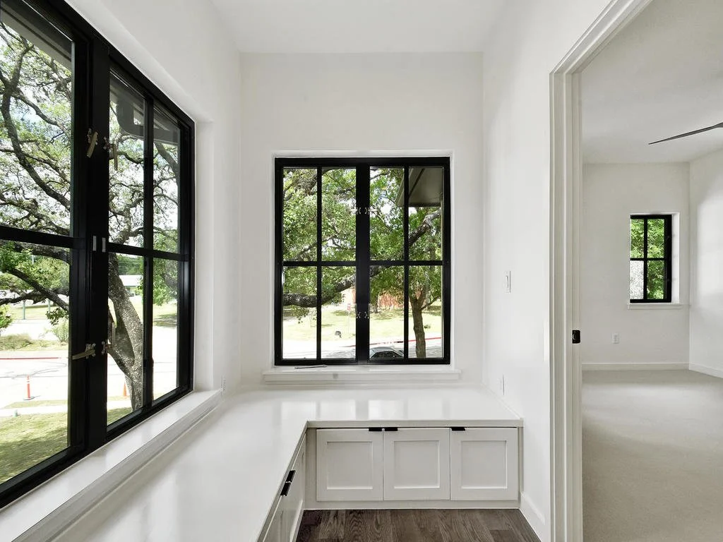 Empty white room with large black-framed windows, built-in storage bench, and view of trees outside.