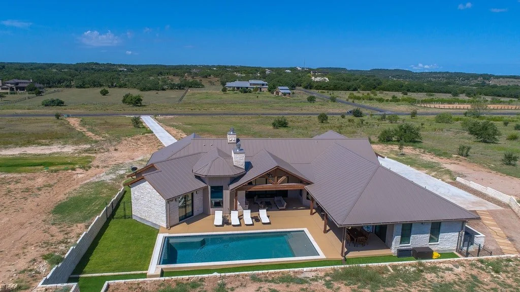 Aerial view of a large modern house with a swimming pool, surrounded by open fields and sparse trees, under a clear blue sky.