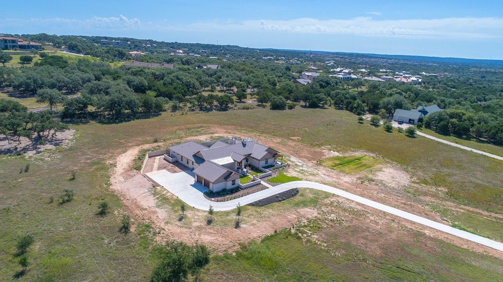 A modern house with a driveway, situated on a large open lot surrounded by trees and open land.