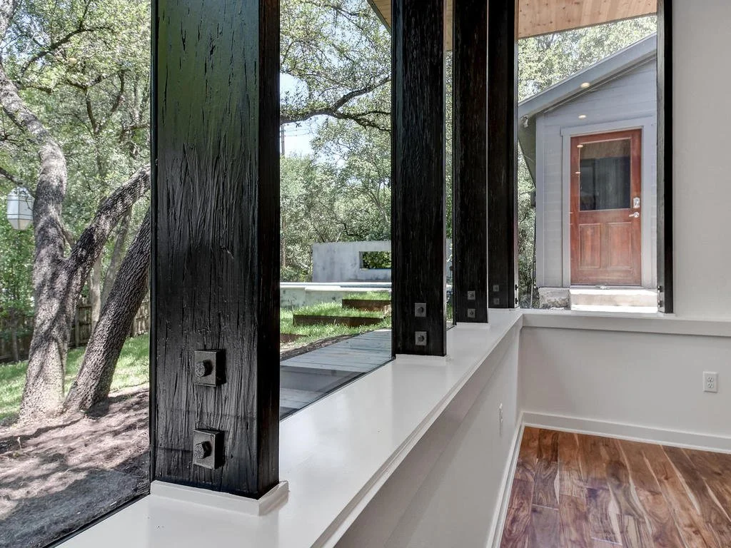 Interior view through large window with black wooden posts, showing a backyard with trees, grass, a patio, and a small gray building with a brown door.