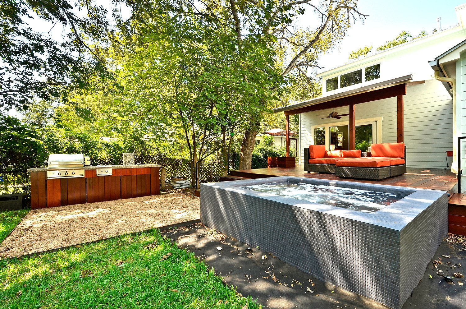 Backyard patio with hot tub, outdoor lounge sofa, small outdoor kitchen with grill, surrounded by trees and greenery.