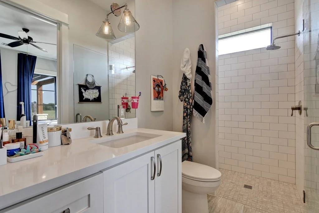 Modern bathroom with a white vanity and sink, large mirror, glass-enclosed shower with white subway tiles, window, and hanging towels.