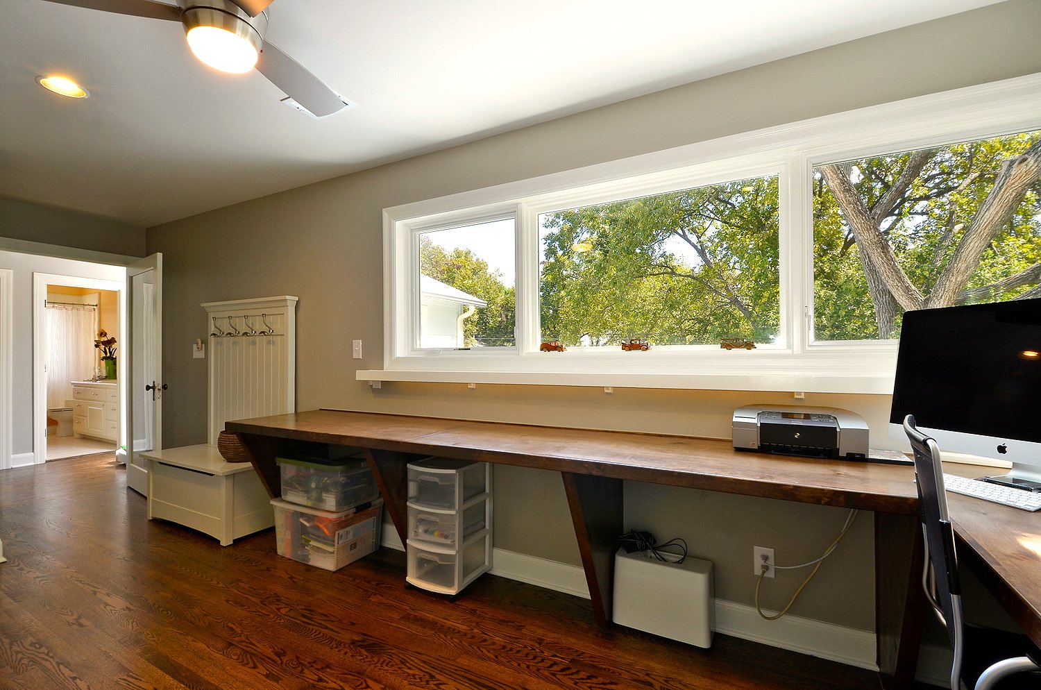 Home office with a long wooden desk, computer, printer, and storage drawers, large window with a view of tree branches outside, hardwood floor, ceiling fan, and a doorway leading to a bathroom.