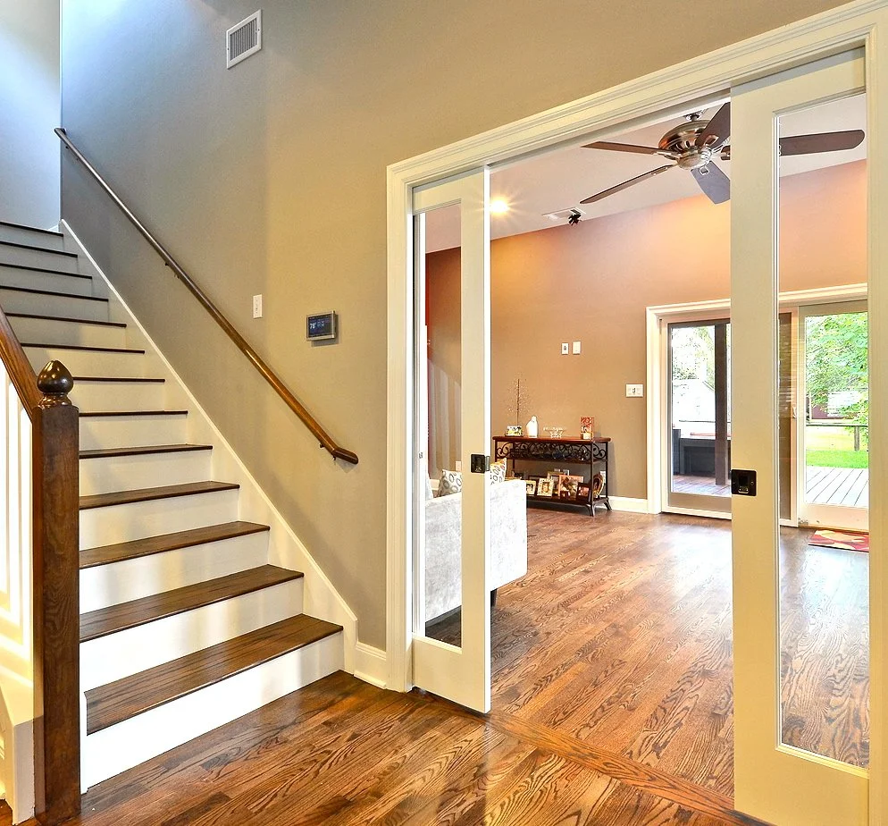 Living room visible through double glass sliding doors, with a ceiling fan, hardwood floors, a window, and a utility shelf with picture frames and decor.