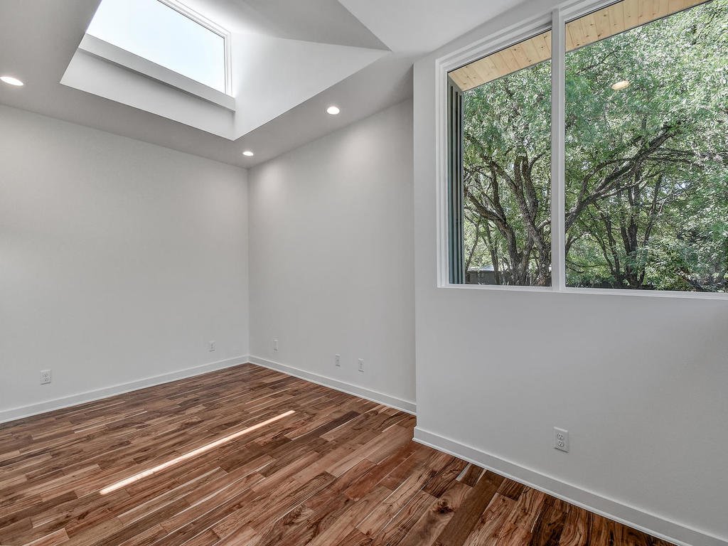 An empty room with white walls, hardwood flooring, a large window showing green foliage outside, and a skylight letting in natural light.