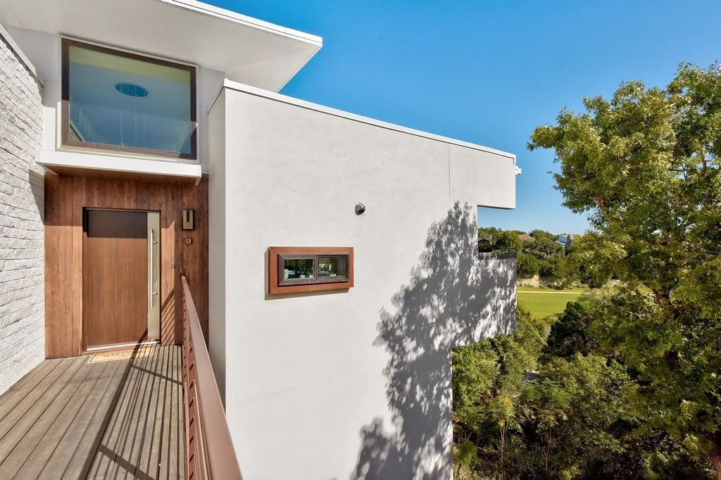 Modern house exterior with a wooden entrance door, a small horizontal window, and a large white wall casting shadows, surrounded by green trees and a bright blue sky.