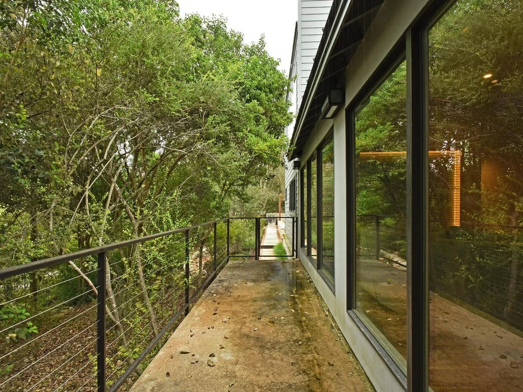 A narrow outdoor balcony with a concrete floor, black railings, and large glass windows reflecting trees, adjacent to a modern building, overlooking a lush wooded area.