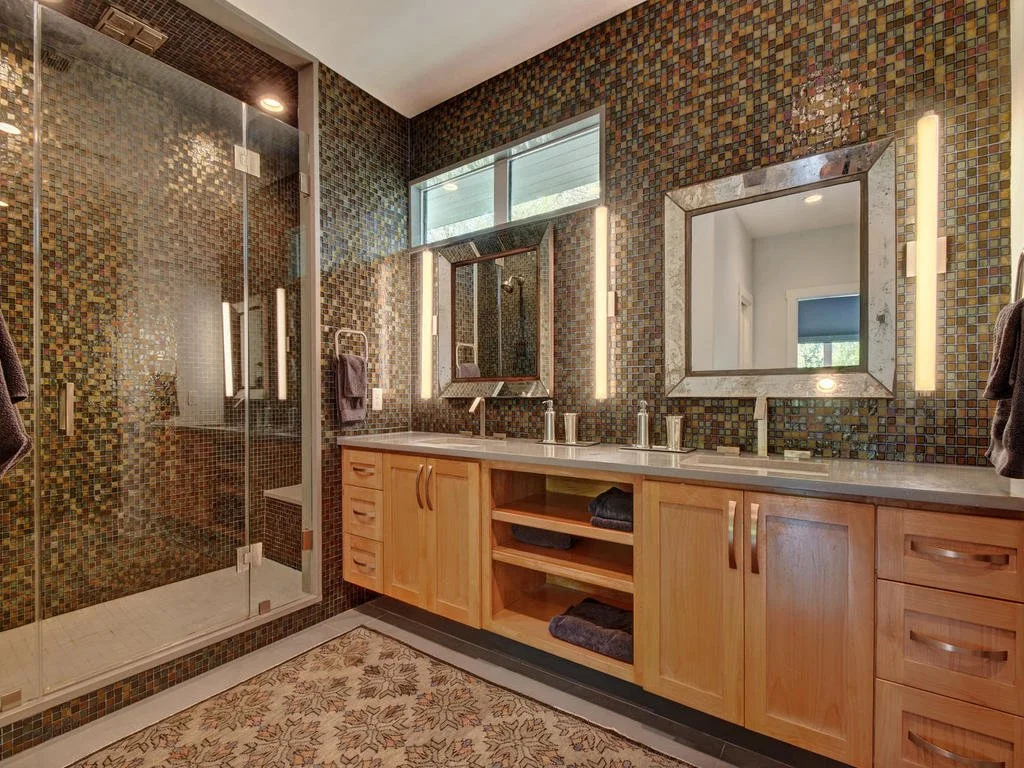 Modern bathroom with mosaic tile walls, a large mirror, and a double sink vanity with wooden cabinets. Includes a walk-in shower with glass doors and a window above the vanity.
