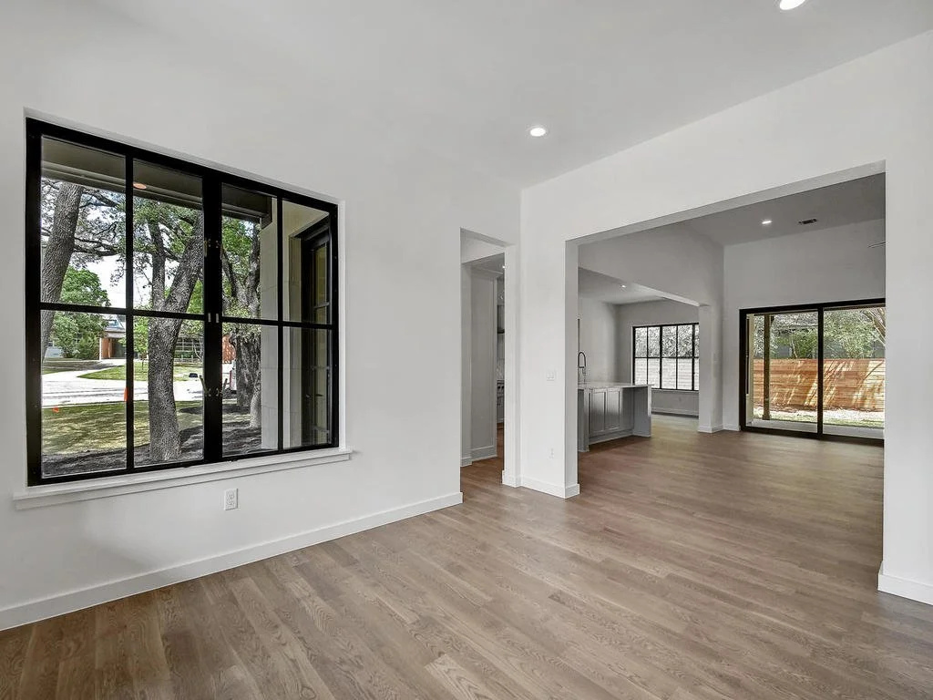 Empty modern living room with white walls, large windows, wood flooring, and an open kitchen in the background.