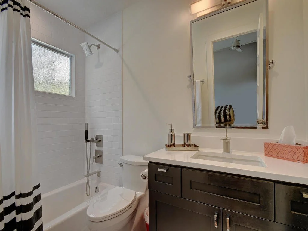 Bathroom with white vanity and black cabinets, white countertop, mirror, tissue box, soap dispenser, toilet, bathtub with shower head, small window, and shower curtain with black and white stripes.