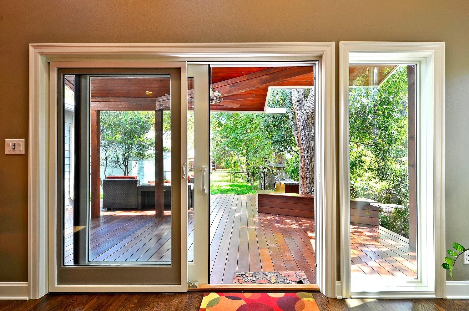Sliding glass door opens to a wooden patio with outdoor furniture and green trees.