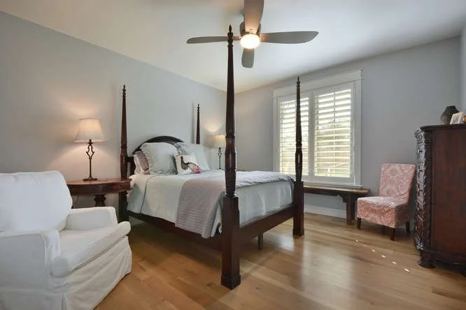 Bedroom with four-poster bed, white bedding, wooden nightstands, a white armchair, pink patterned armchair, wooden dresser, window with white shutters, ceiling fan, hardwood flooring.