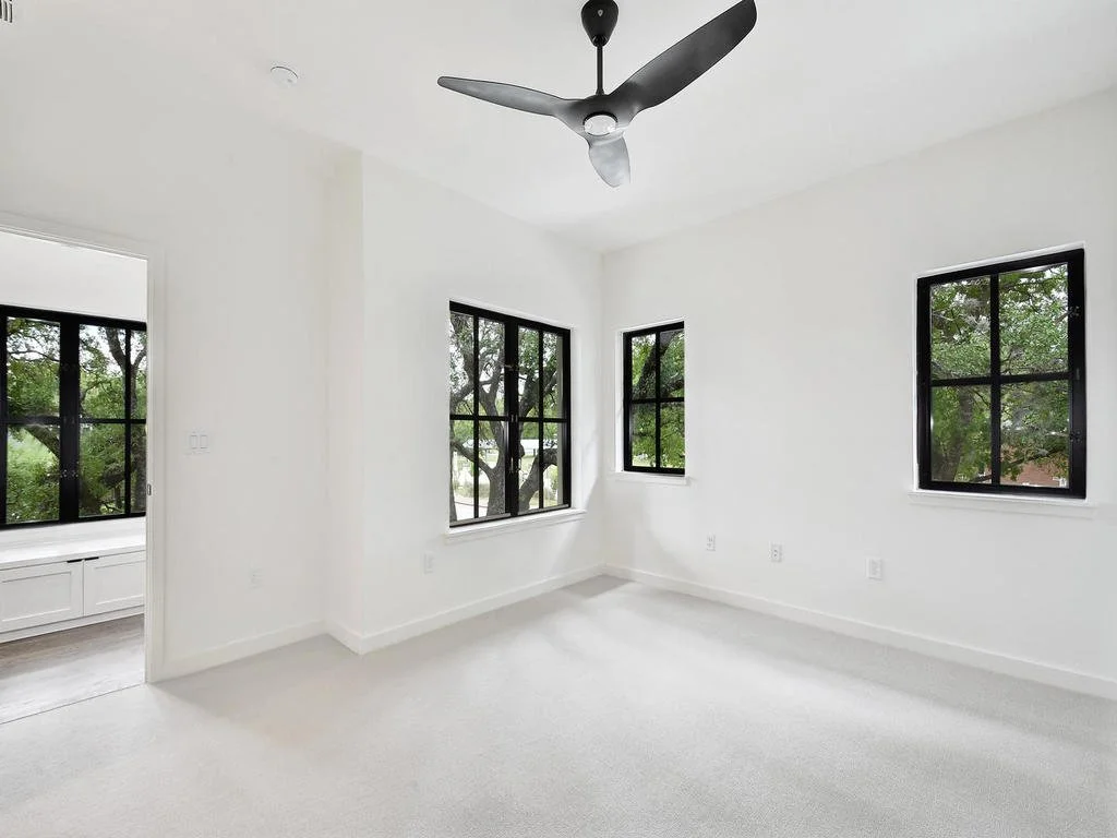 Empty room with white walls and ceiling, black-framed windows showing trees outside, a ceiling fan, and a light-colored carpet.