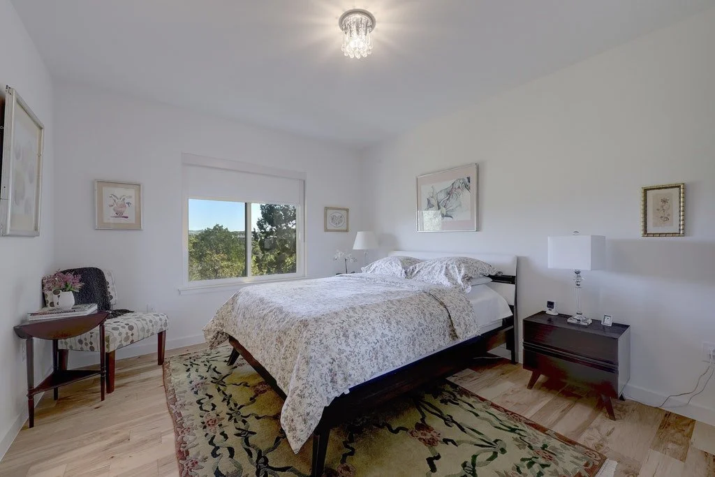 A bedroom with white walls, a bed with patterned bedding, a dark wooden nightstand with a white lamp, a decorative rug, a window showing greenery outside, and framed artwork on the walls.