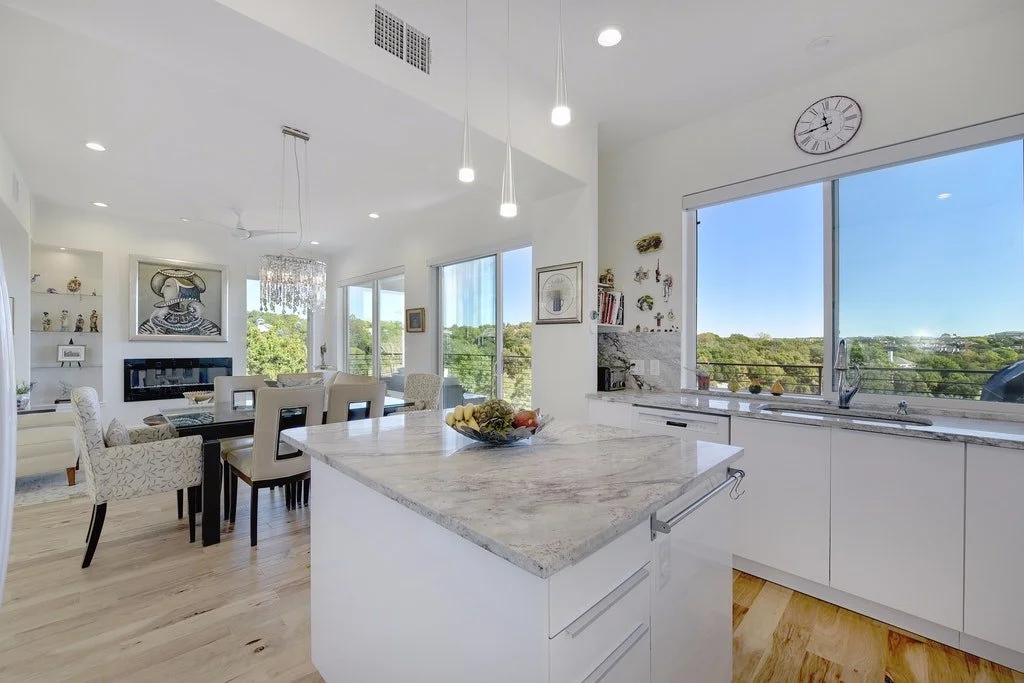 Open-concept modern kitchen and dining area with large windows, white cabinets, light wood flooring, and minimalistic decor.