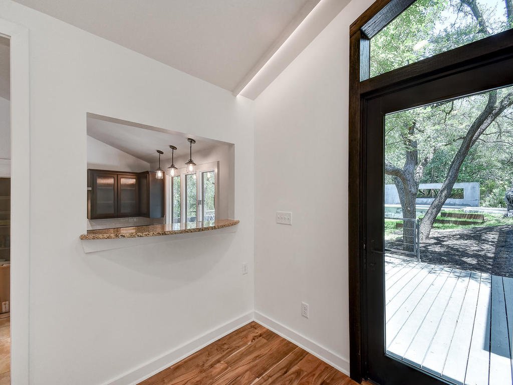 Interior view of a house with a doorway leading to a deck outside, showing a kitchen with pendant lights and windows overlooking trees.