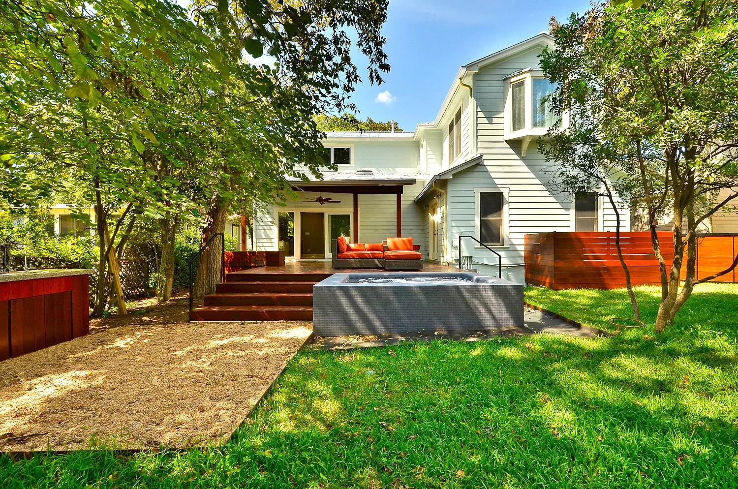 Backyard of a white house with a wooden deck, orange outdoor furniture, green lawn, trees, and a hot tub.