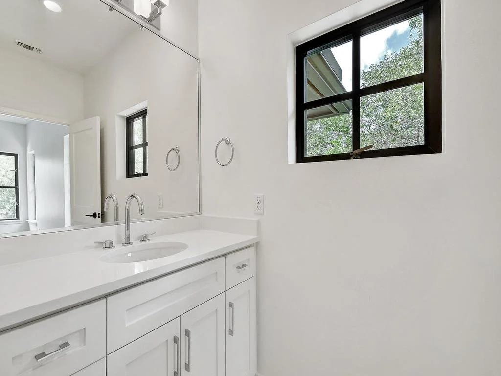 A clean, modern bathroom vanity with a white countertop, a silver faucet, and cabinet doors, with a large mirror above, a towel ring, and a window showing outside greenery.