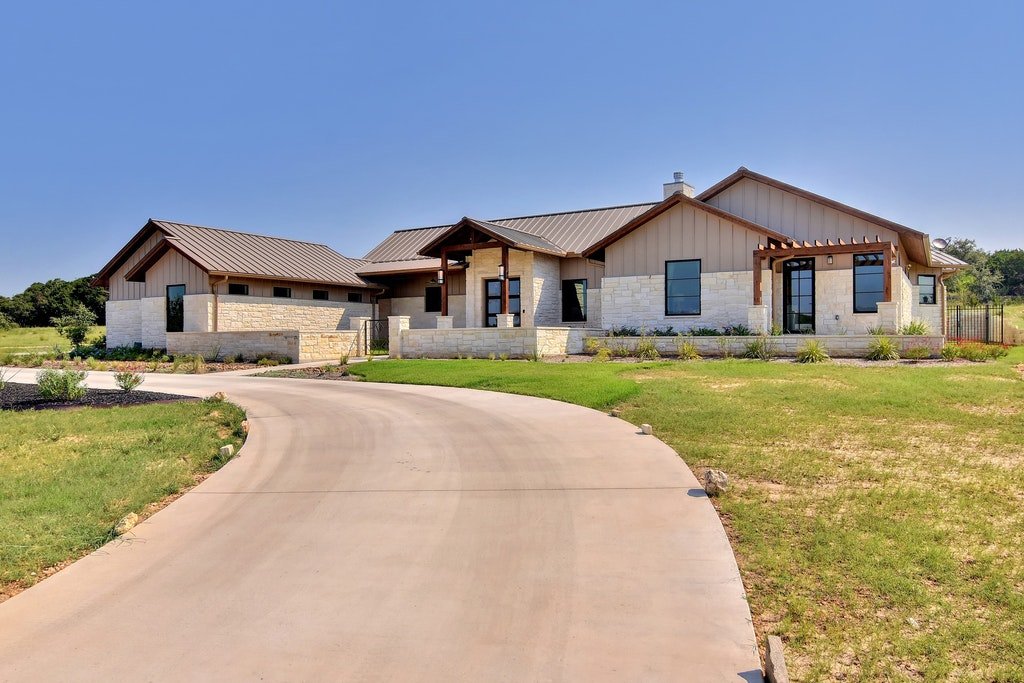 New modern house with a circular driveway, stone and wood exterior, large windows, and landscaped yard under a clear blue sky.