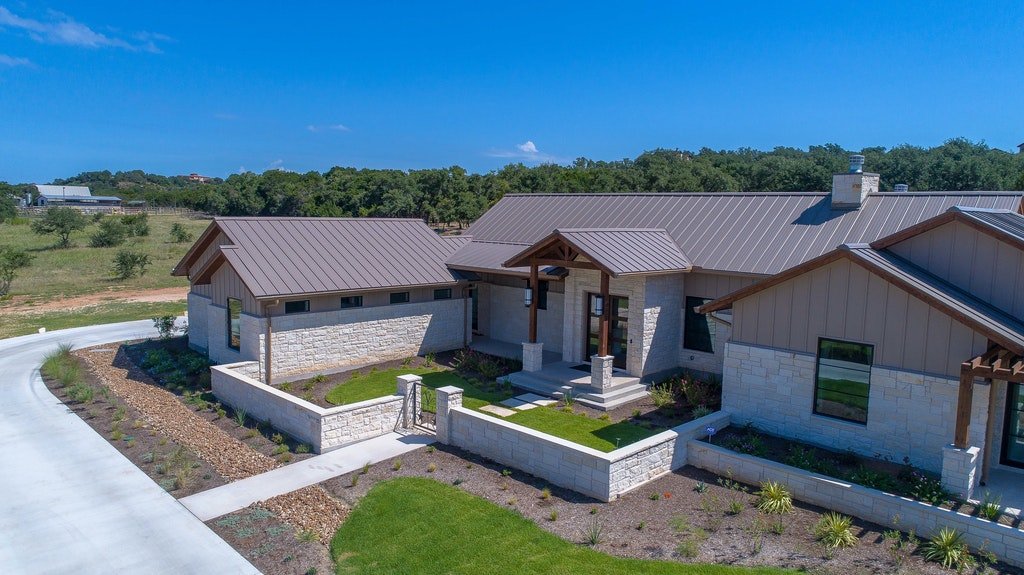 A modern single-story house with stone and wood exterior, metal roof, front porch, and landscaped yard, situated on a curved driveway in a rural area with trees in the background.