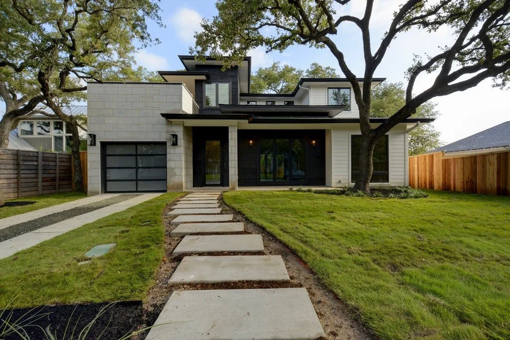 Modern two-story house with a gray and white exterior, large front windows, and a concrete walkway leading to the front door, with a well-maintained grassy lawn and trees in the yard.
