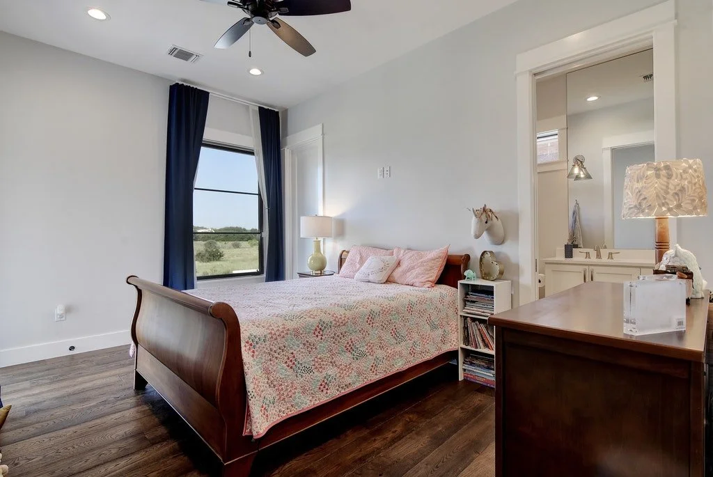 A bedroom with a wooden bed featuring pink and floral bedding, a white nightstand with books and decor, a lamp, a window with navy curtains, and an attached bathroom visible through an open door.