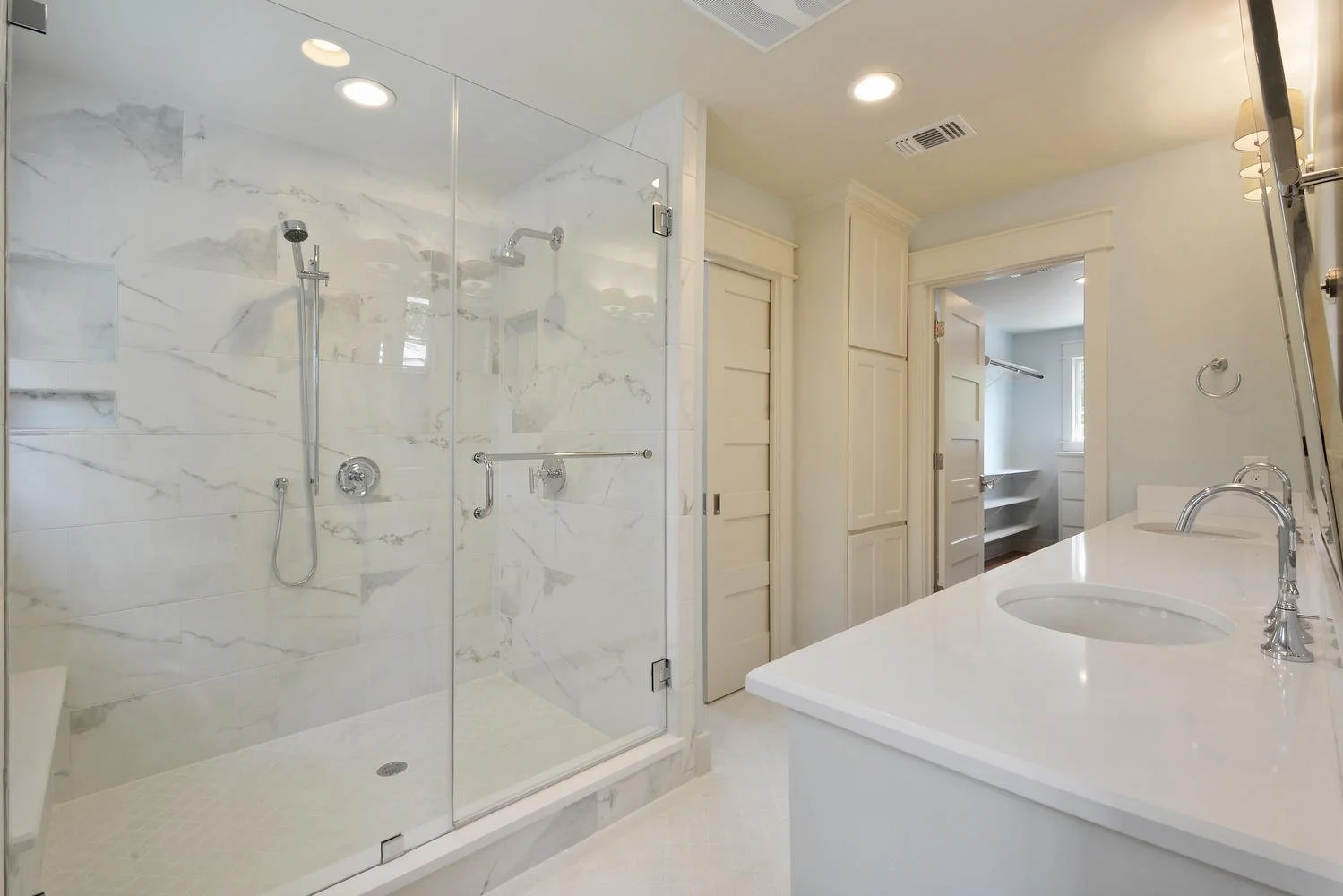 Modern bathroom with glass-enclosed shower featuring marble walls, a white vanity with dual sinks, and a walk-in pantry visible through a door