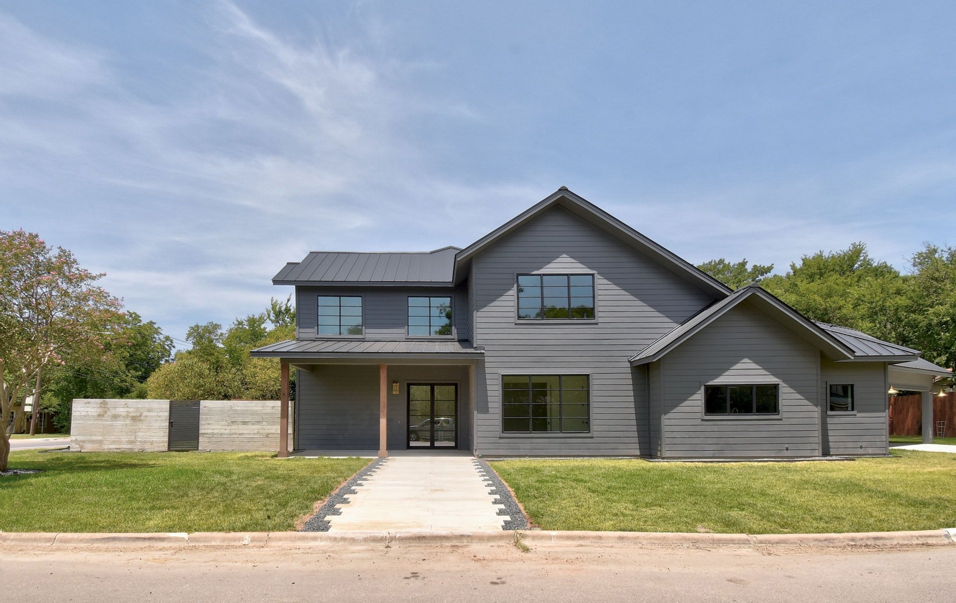 A modern two-story gray house with multiple windows, a metal roof, and a front porch. The house is surrounded by a green lawn, trees, and a concrete sidewalk leading to the entrance. The sky is partly cloudy.