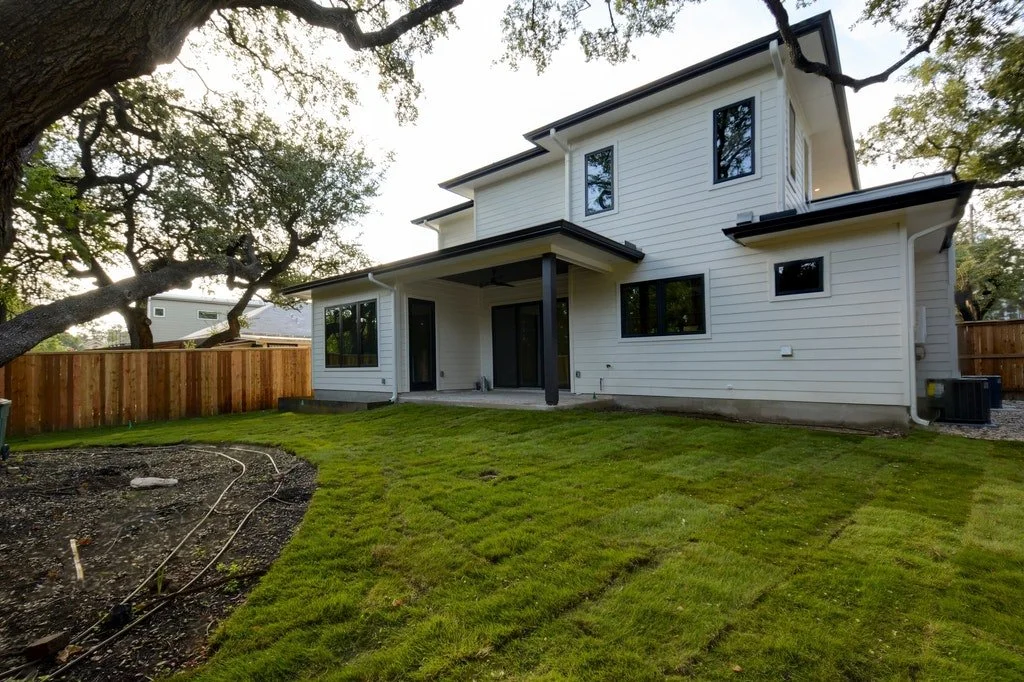 A two-story white house with black trim, multiple windows, and a covered back porch with a black support column. The backyard has a well-maintained green lawn, a wooden privacy fence, and some trees providing shade.